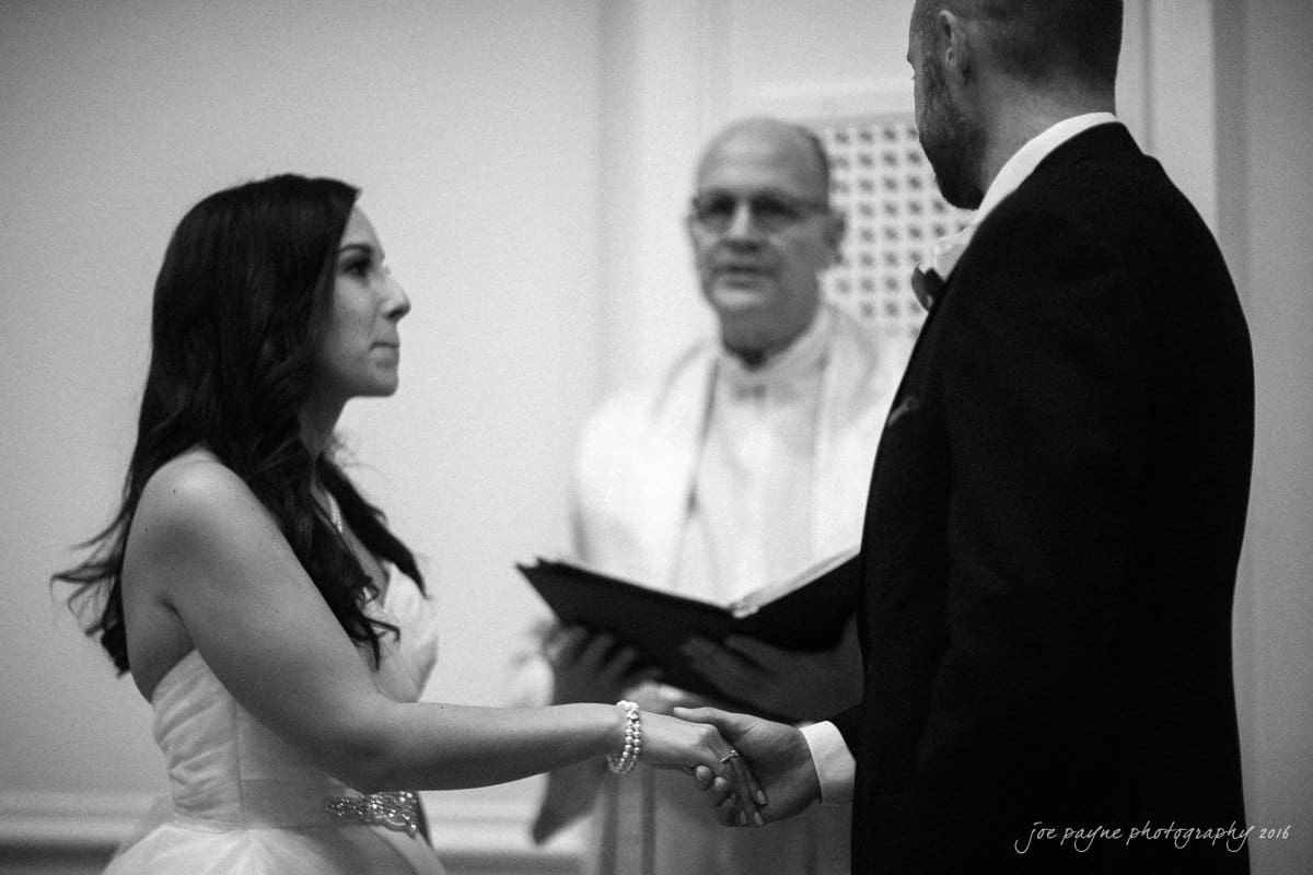 carolina inn wedding photography - kristen & matthew (x2) A bride and groom hold hands during a wedding ceremony. The officiant, wearing glasses and holding a book, stands in the background. The black and white image captures a solemn moment between the couple.