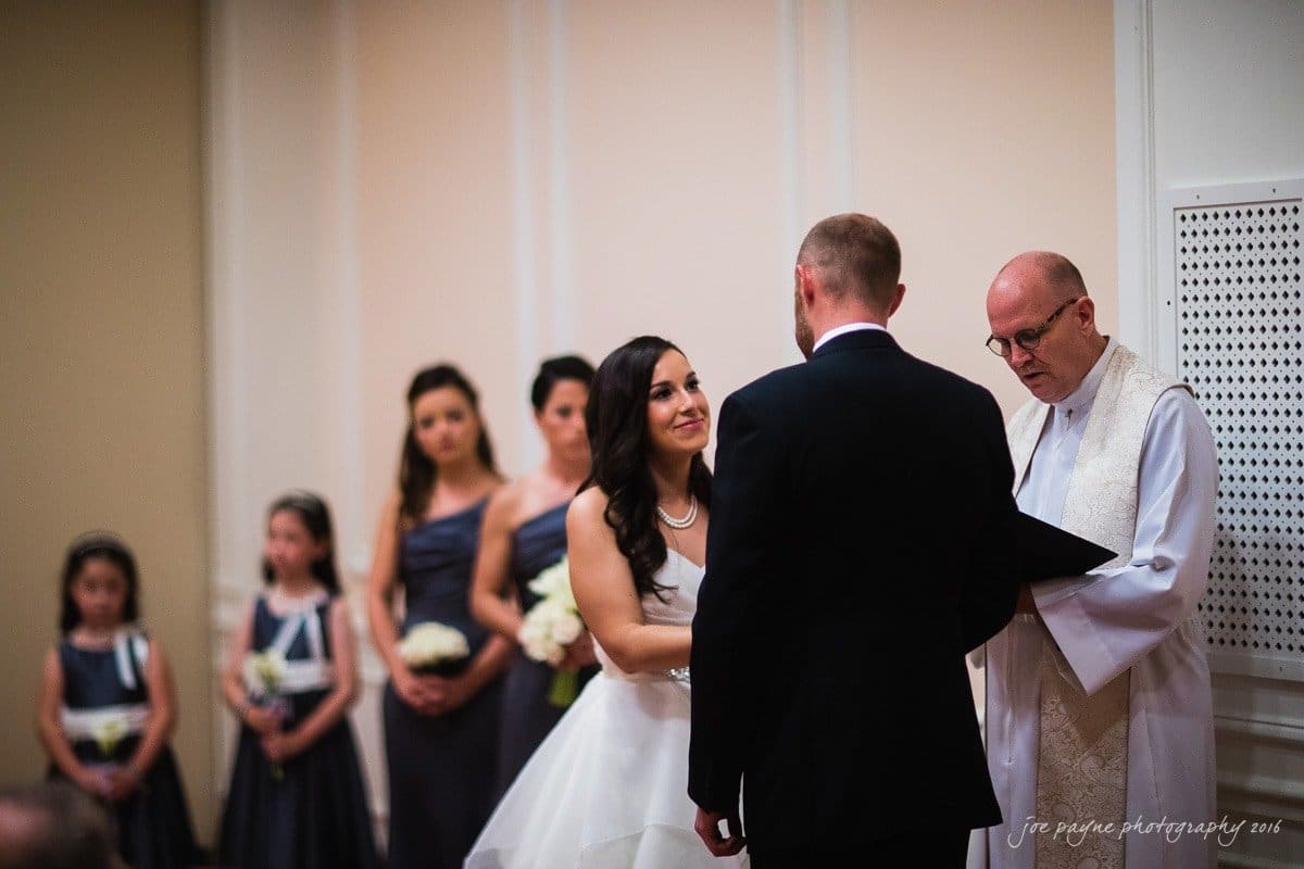 carolina inn wedding photography - kristen & matthew (x2) A bride and groom stand facing each other during their wedding ceremony. A minister, holding a book, stands beside them. Bridesmaids and flower girls in matching dresses watch from the side.
