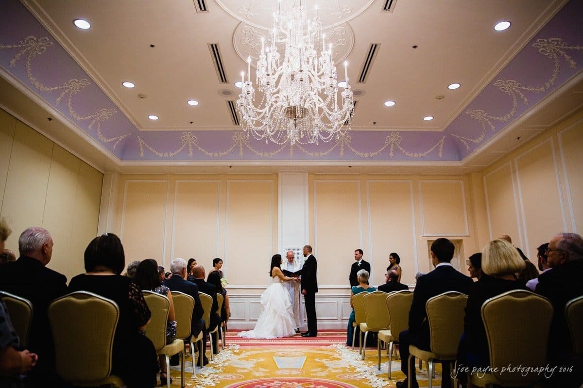 carolina inn wedding photography - kristen & matthew (x2) A couple stands at the altar in an elegant room with a large chandelier above. Guests are seated on both sides, witnessing the ceremony. The room is softly lit, with light purple and cream walls and ornate ceiling details.
