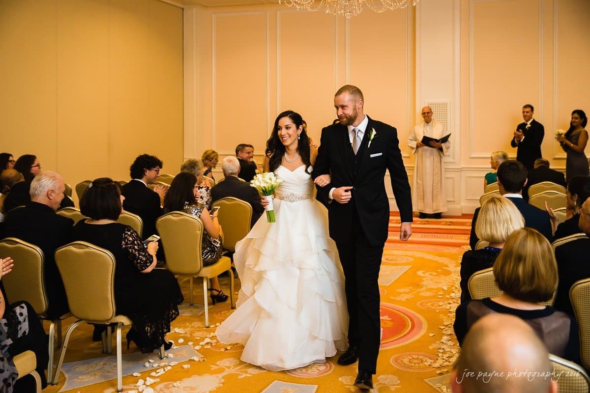 carolina inn wedding photography - kristen & matthew (x2) A bride in a white gown and a groom in a black suit walk arm-in-arm down the aisle in a wedding ceremony. Guests seated on either side applaud. A priest stands in the background. The room is elegantly decorated with chandeliers.