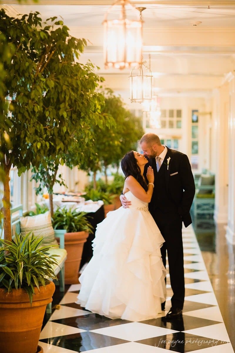 carolina inn wedding photography - kristen & matthew (x2) A couple stands in a bright hallway with black and white checkered floors. The woman in a white gown holds the mans face, who is in a black suit. They are surrounded by potted plants and soft lighting from overhead lanterns.