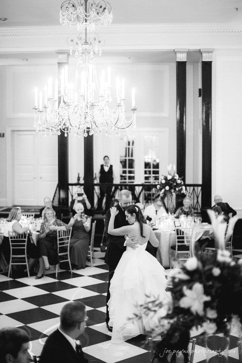 carolina inn wedding photography - kristen & matthew (x2) A black and white photo of a bride and groom dancing on a checkered floor in an elegant venue. A chandelier hangs above, and guests sit at round tables watching the couple. Flowers and candles decorate the room, and a person stands in the background.