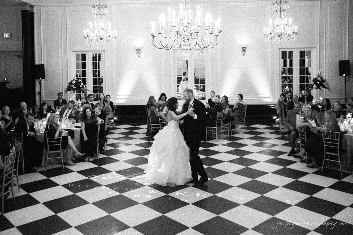 carolina inn wedding photography - kristen & matthew (x2) A bride and groom dance in a grand ballroom with checkered floors. Guests are seated around them at tables. Elegant chandeliers hang from the ceiling, and soft lighting adds to the romantic atmosphere.
