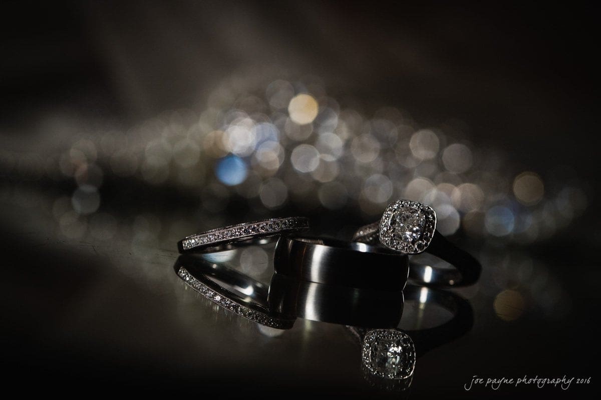 carolina inn wedding photography - kristen & matthew (x2) A close-up of three rings on a reflective surface with a blurred, sparkling background. Two rings are simple bands, and the third features a central, square-set gem surrounded by smaller stones.