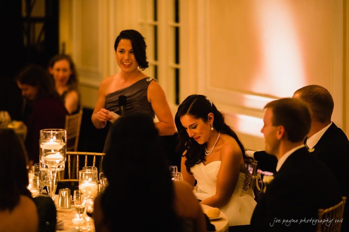 carolina inn wedding photography - kristen & matthew (x2) A woman in a sleeveless dress holds a microphone and speaks at a formal event. A seated woman in a white gown smiles while listening. Guests are seated at tables decorated with candles and glasses in a warmly lit room.