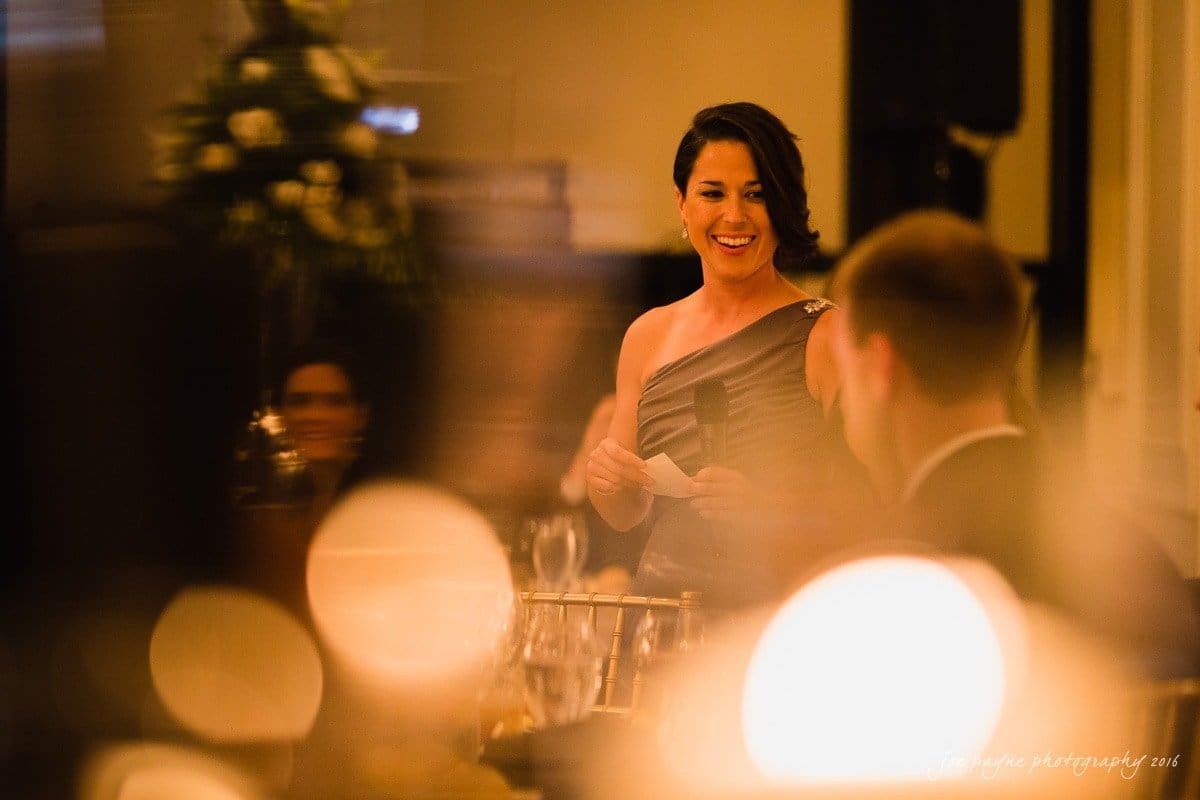 carolina inn wedding photography - kristen & matthew (x2) A woman in an elegant off-shoulder dress is speaking joyfully at an event. The scene is warmly lit with blurred lights and guests in the background, creating a festive ambiance.