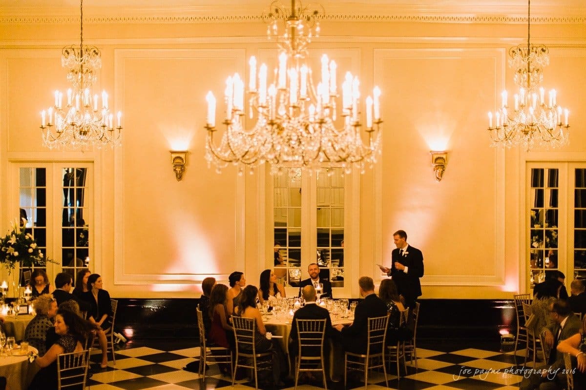 carolina inn wedding photography - kristen & matthew (x2) A warm, elegant ballroom scene with guests seated at round tables under ornate chandeliers. A person stands giving a speech, while others listen attentively. The room features high ceilings, large mirrors, and soft lighting.