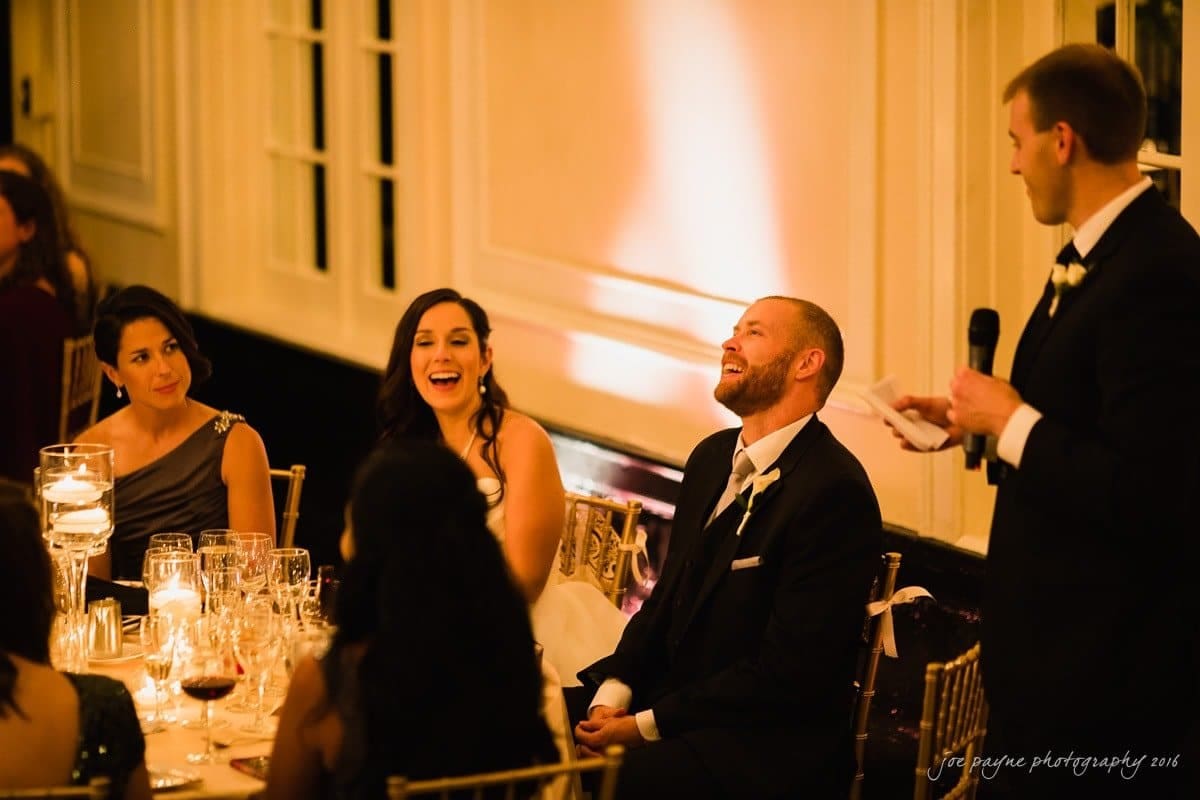 carolina inn wedding photography - kristen & matthew (x2) A man stands and speaks into a microphone, smiling, while three seated guests at a formal event laugh and listen. The room is warmly lit, with elegant table settings and candles.