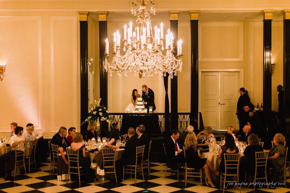 carolina inn wedding photography - kristen & matthew (x2) A dimly lit ballroom with a large chandelier. A couple stands at a table, cutting a cake. Guests are seated at round tables with white tablecloths and golden chairs, engaged in conversation. Black and white checkered floor completes the elegant setting.