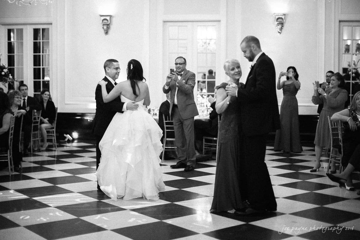 carolina inn wedding photography - kristen & matthew (x2) A wedding reception with two couples dancing on a checkered floor. The bride and groom are in the foreground, while a man and an older woman dance nearby. Guests sit and stand around, watching and capturing the moment with their phones.