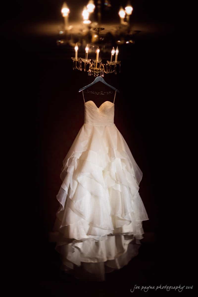 carolina inn wedding photography - kristen & matthew (x2) A white strapless wedding gown with layered, ruffled fabric is elegantly hung on a hanger under a lit chandelier in a dimly lit room. The soft light highlights the dresss texture and design.