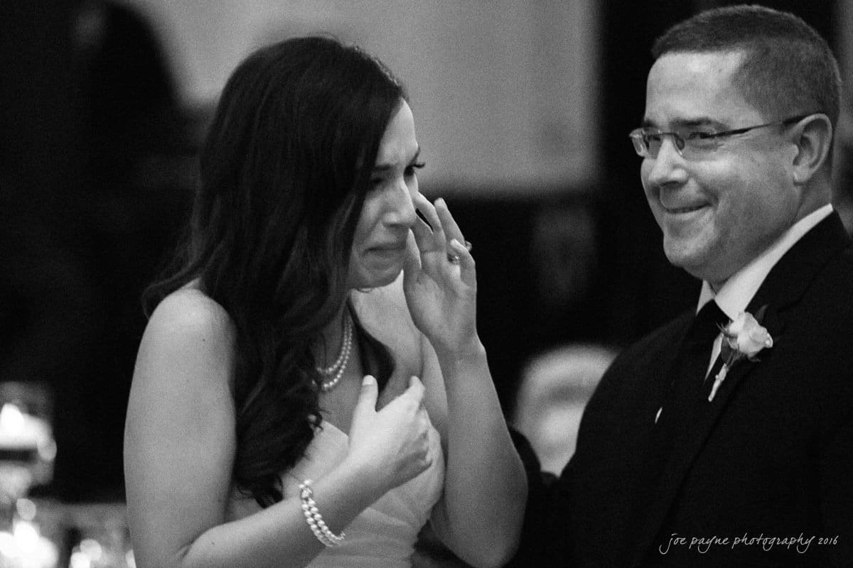 carolina inn wedding photography - kristen & matthew (x2) A bride wipes tears from her eyes as she smiles during a dance with an older man in a suit and glasses at a wedding reception. Both appear emotional and joyful. The background is softly blurred.