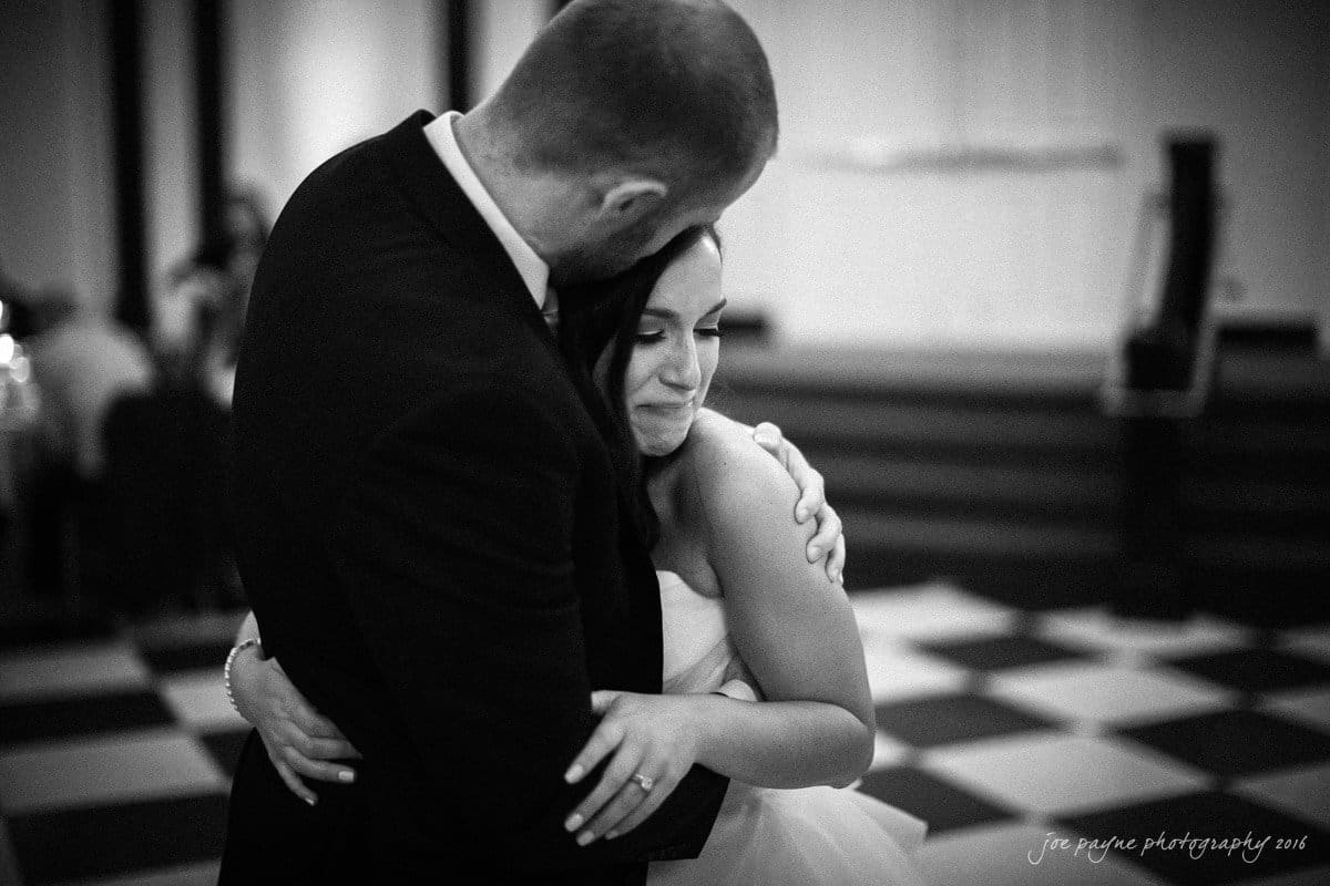 carolina inn wedding photography - kristen & matthew (x2) A bride and groom share an intimate dance on a checkered floor. The groom is in a black suit holding the bride, who wears a white dress, as she rests her head against his chest with a content expression. The setting is softly lit and elegant.