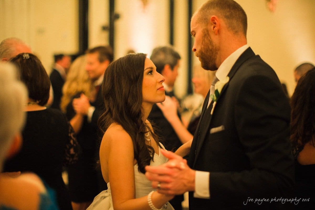 carolina inn wedding photography - kristen & matthew (x2) A couple dances closely at a formal event. The woman in a white dress looks up at the man in a suit. The background is softly blurred with warm lighting and other guests mingling.