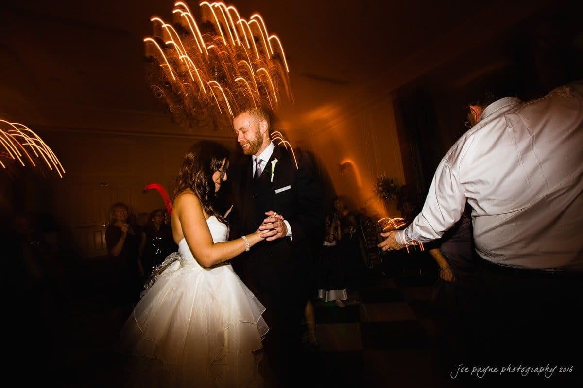 carolina inn wedding photography - kristen & matthew (x2) A bride and groom share a dance at their wedding reception. The room is dimly lit with streaks of light captured in motion. The groom wears a suit and the bride a white gown as they dance surrounded by guests.