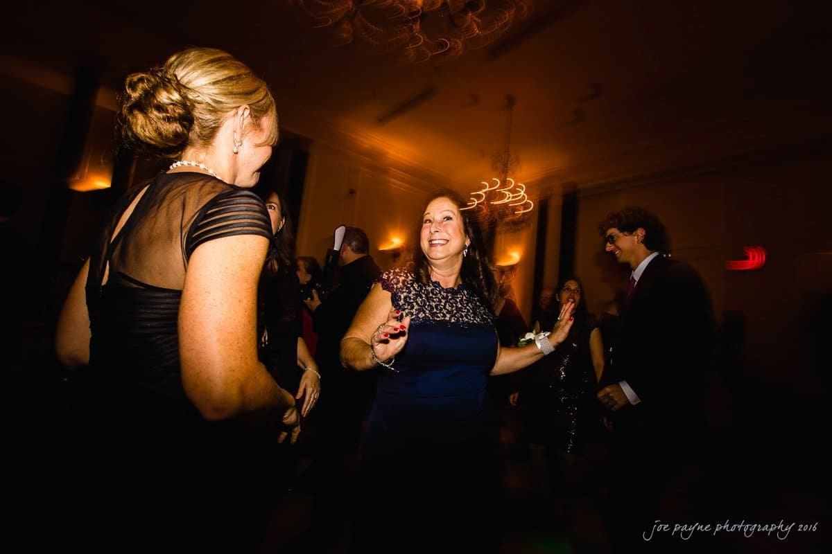 carolina inn wedding photography - kristen & matthew (x2) A group of people dance and laugh in a dimly lit room. The woman in the center, wearing a blue dress, smiles joyfully. Others around her are dressed formally. The ambiance is festive with soft warm lighting.