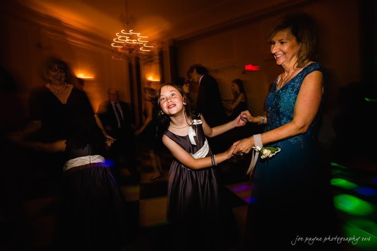 carolina inn wedding photography - kristen & matthew (x2) A young girl and an older woman joyfully dance together in a dimly lit room. The girl is wearing a black dress with a white sash, while the woman is in a blue dress. Other people are dancing in the background, and colorful lights illuminate the floor.