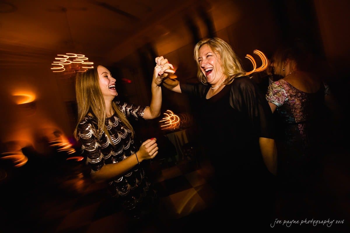carolina inn wedding photography - kristen & matthew (x2) Two women joyfully dance together at a party. One wears a patterned dress, the other a black outfit. They smile broadly, surrounded by blurred lights and a lively atmosphere, conveying a sense of fun and celebration.
