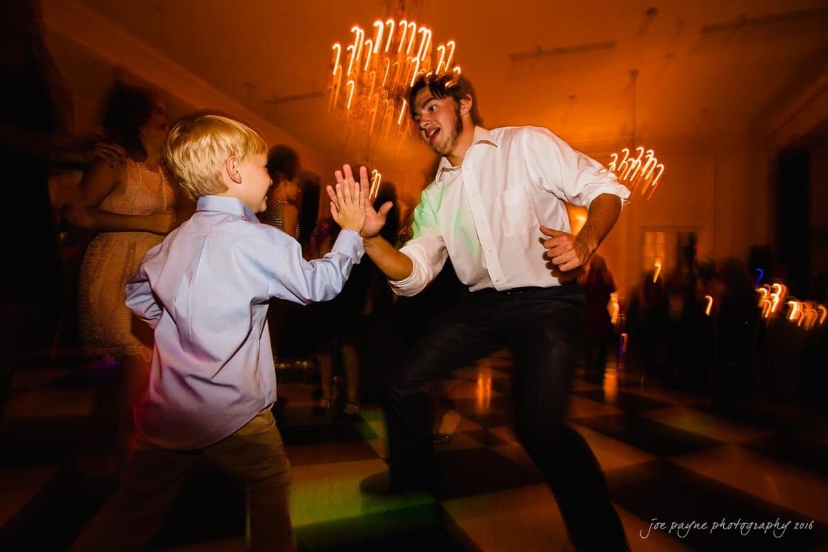 carolina inn wedding photography - kristen & matthew (x2) A young boy and a man in a white shirt high-five each other enthusiastically on a dance floor. The background is colorful with blurred lights and people. The atmosphere is lively and joyful.