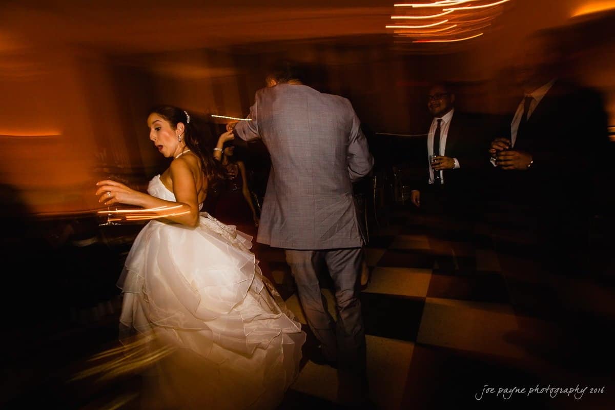 carolina inn wedding photography - kristen & matthew (x2) A bride in a white gown dances with a man in a gray suit on a checkered floor. The blurred motion and warm lighting create a lively atmosphere. Other guests are visible in the background holding drinks.