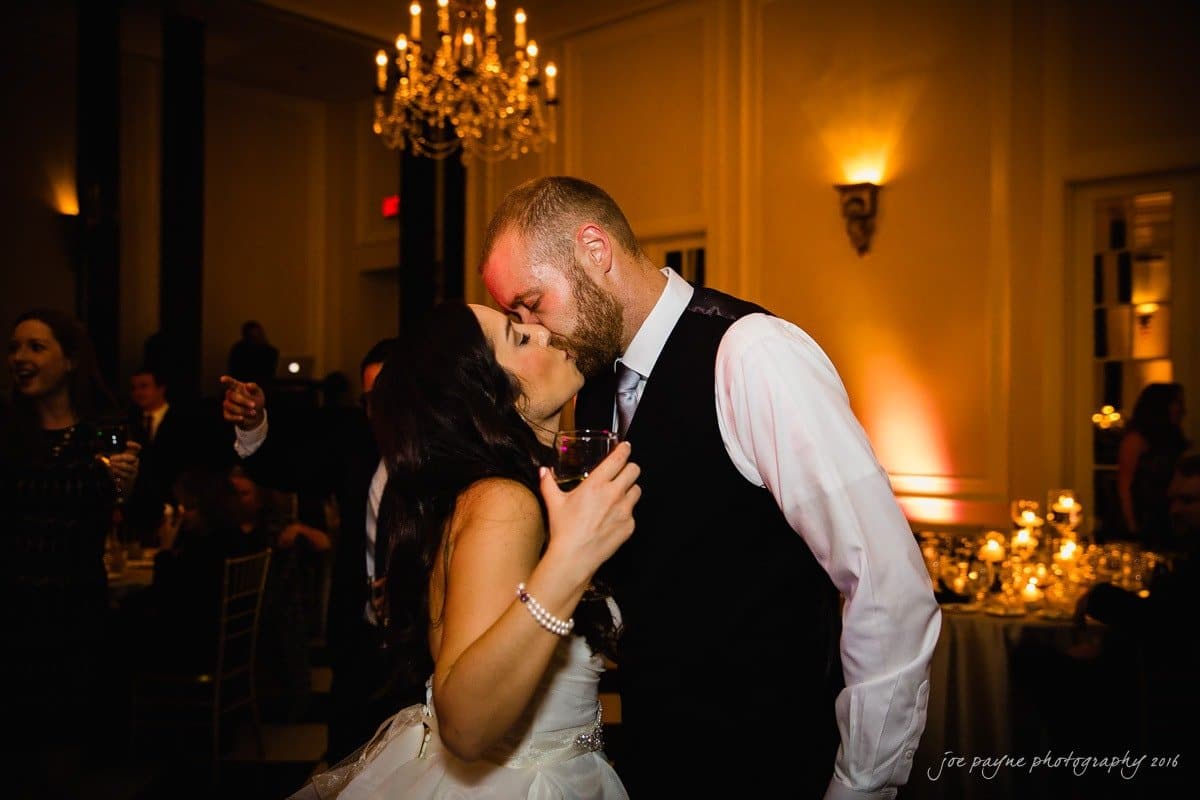 carolina inn wedding photography - kristen & matthew (x2) A bride and groom share a kiss in a warmly lit ballroom. The bride holds a glass of wine, and both are dressed elegantly. A chandelier hangs above them, and guests are visible in the background.