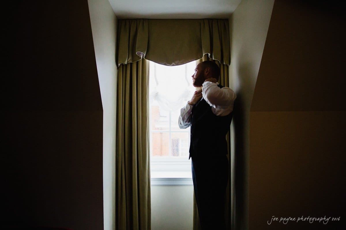 carolina inn wedding photography - kristen & matthew (x2) A man stands in profile, adjusting his tie, silhouetted against a bright window with draped curtains. The room is dimly lit, creating a strong contrast with the light streaming in.