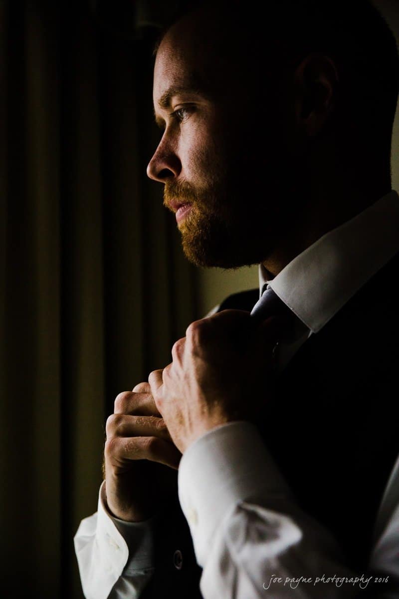 carolina inn wedding photography - kristen & matthew (x2) A man with a beard is adjusting his tie while looking to the side. He is wearing a suit and a white shirt. The lighting highlights one side of his face, creating a dramatic shadow effect.