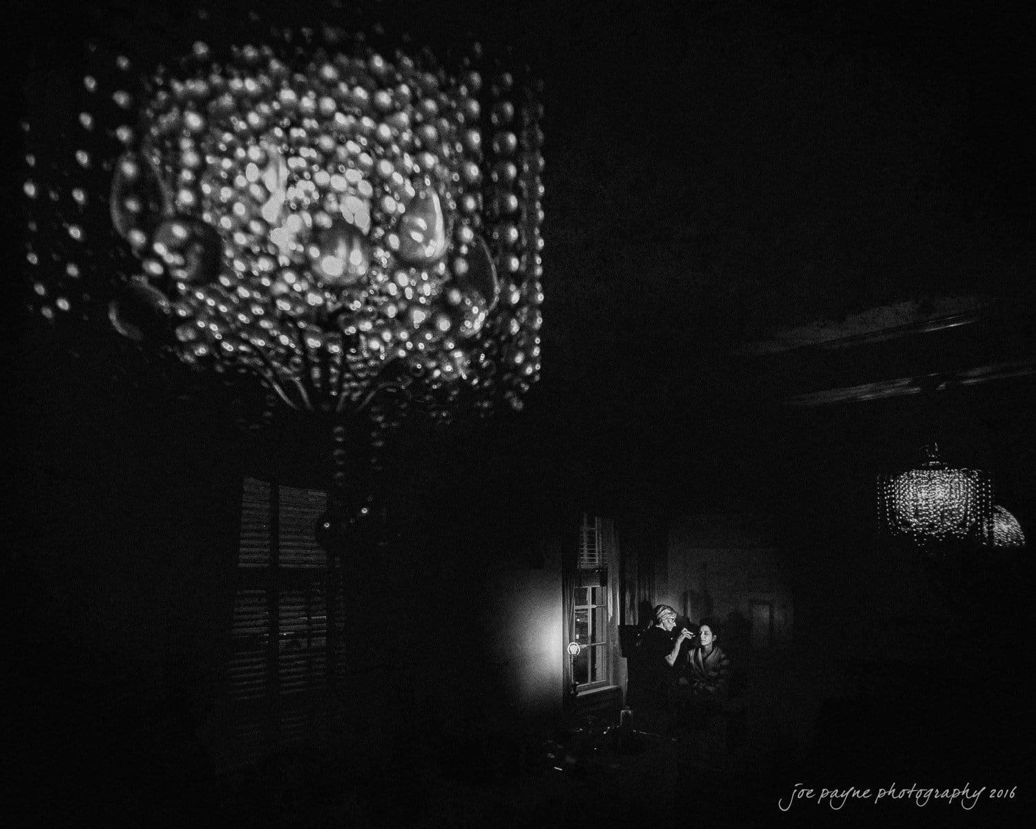 carolina inn wedding photography - kristen & matthew (x2) A black and white photo of a dimly lit room with an illuminated chandelier reflecting on the wall. Two people, one in a white outfit, are sitting near a window, engaged in conversation. The ambiance is intimate and cozy.