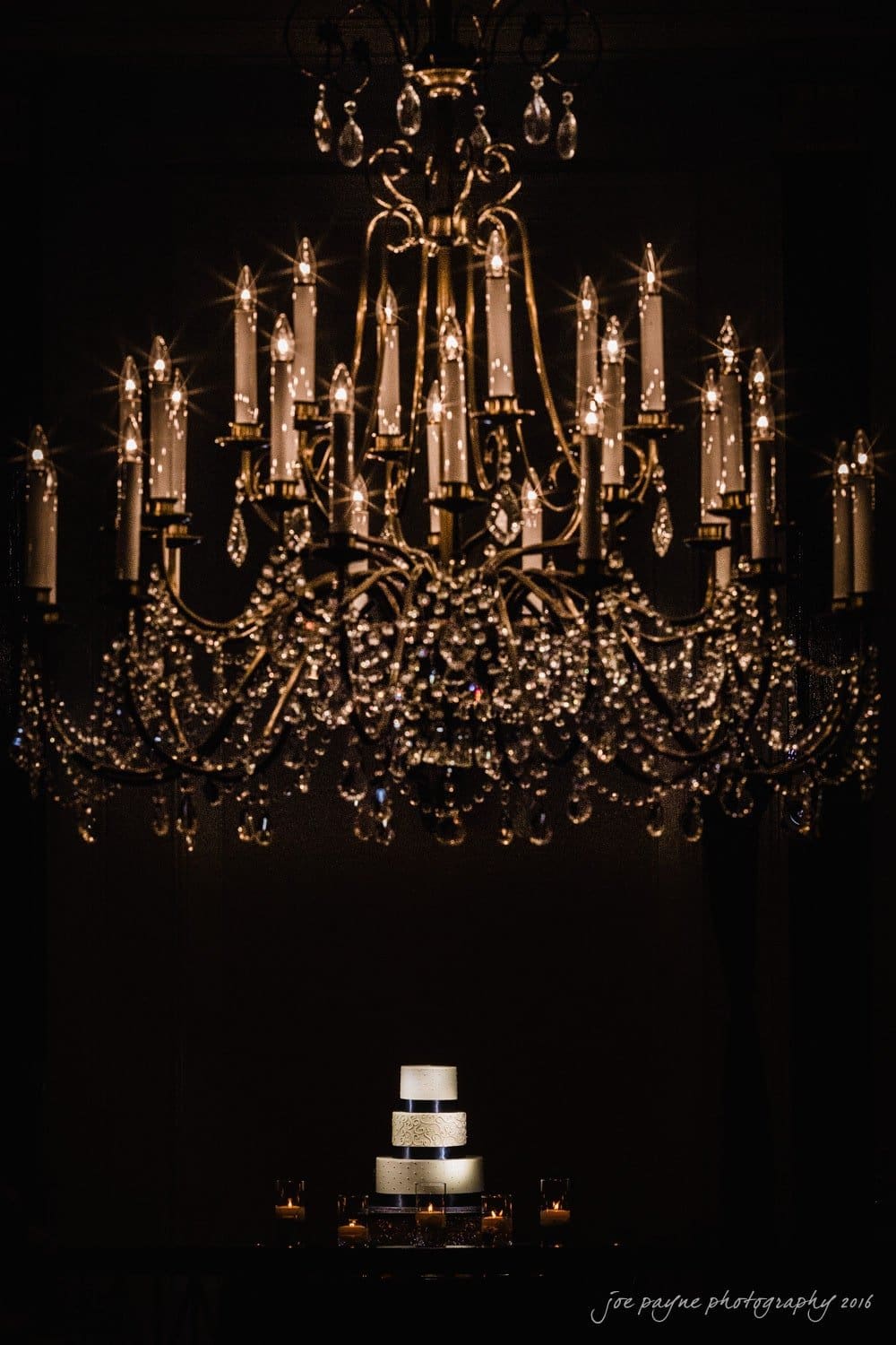 carolina inn wedding photography - kristen & matthew (x2) A large, elegant chandelier is illuminated with many small lights, casting a soft glow over a tiered white wedding cake below. The scene is dark, accentuating the brilliance of the chandelier and the cake.