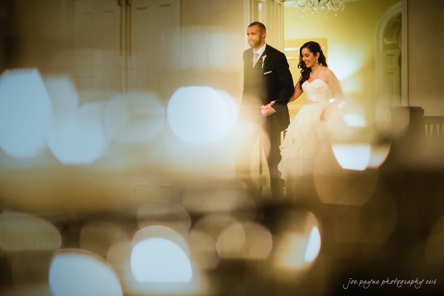 carolina inn wedding photography - kristen & matthew (x2) A bride and groom stand together, holding hands at an elegant indoor venue. The foreground is blurred with ambient lights, creating a warm and romantic atmosphere. The bride wears a gown, and the groom is in a suit.