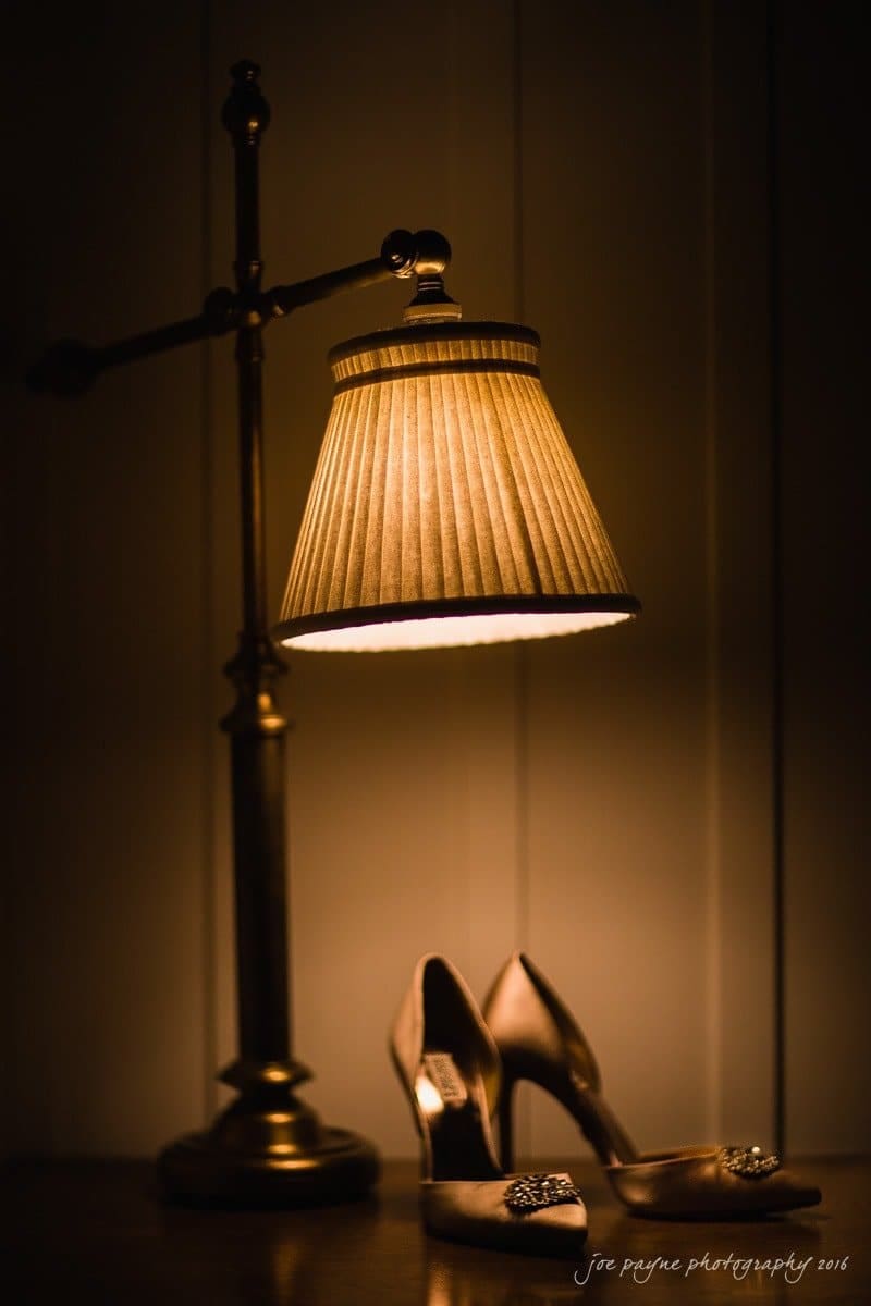 carolina inn wedding photography - kristen & matthew (x2) A pair of elegant high-heeled shoes is placed under a vintage brass lamp with a pleated lampshade, casting a warm, soft glow in a dimly lit setting.