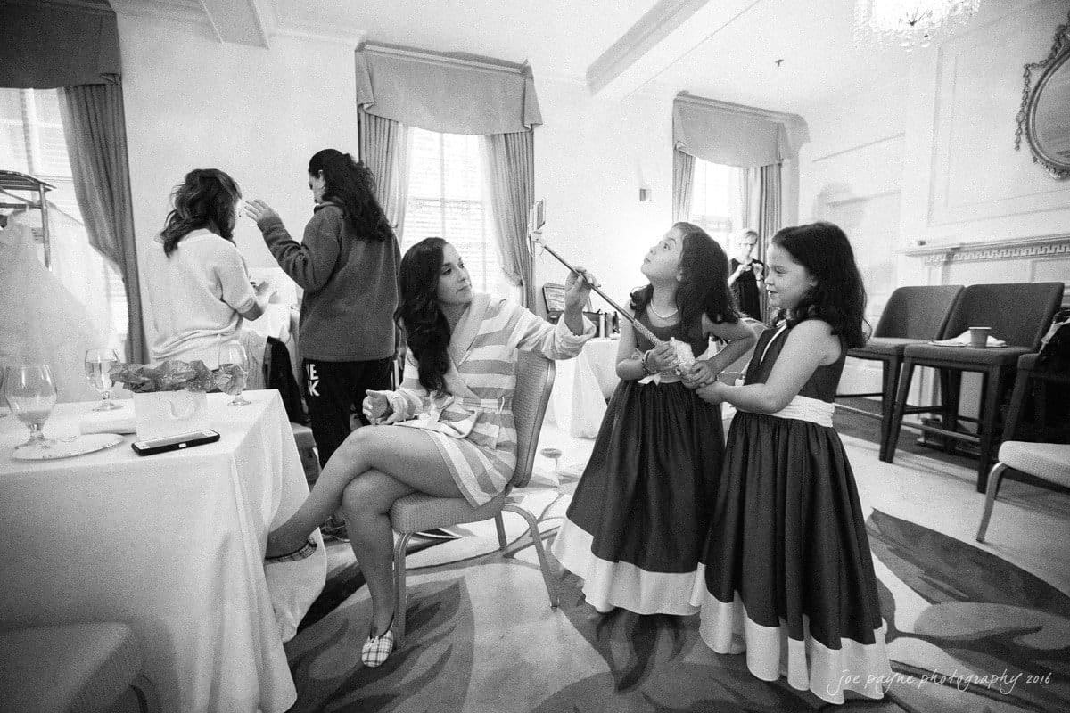 carolina inn wedding photography - kristen & matthew (x2) A woman takes a selfie with two young girls in matching dresses. The room is elegantly furnished, with people in the background preparing and interacting. Natural light streams through large windows, creating a warm, lively atmosphere.