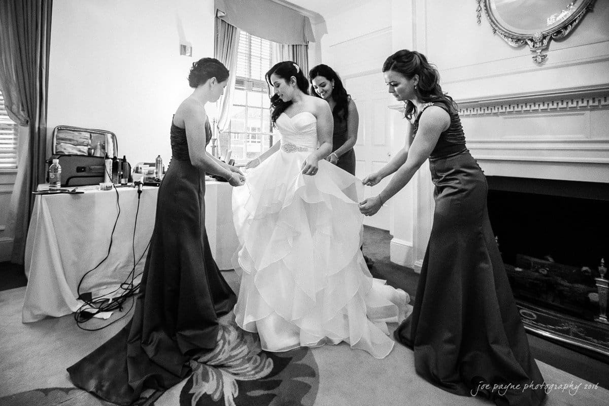 carolina inn wedding photography - kristen & matthew (x2) A bride in a white gown is surrounded by three bridesmaids in dark dresses, assisting with her dress. They are in a elegantly decorated room with a fireplace and a mirror.