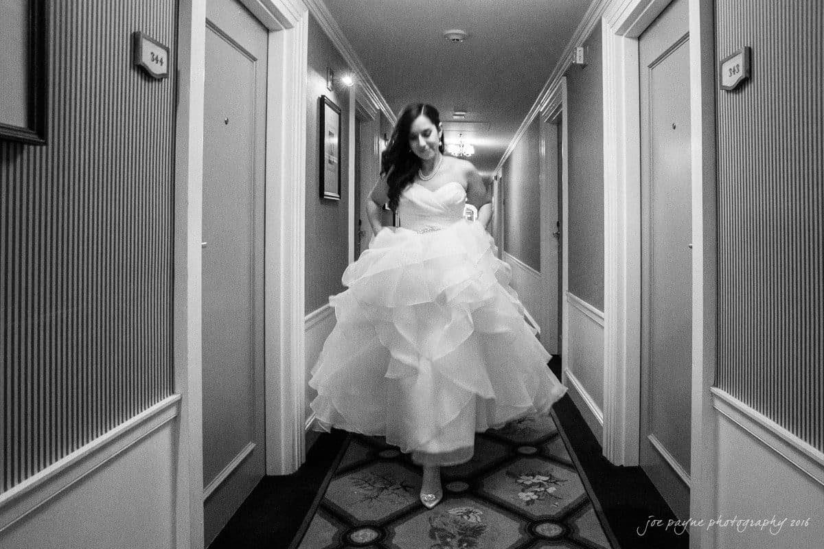 carolina inn wedding photography - kristen & matthew (x2) Bride in a flowing gown joyfully walking down a hotel hallway. The corridor features patterned carpets and framed pictures on the walls, with numbered doors on either side. The image is in black and white.