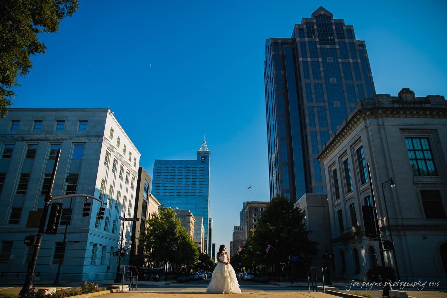 carolina inn wedding photography - kristen & matthew (x2) A person in a formal dress stands in the middle of a city street, surrounded by tall buildings under a clear blue sky. The sun casts long shadows, highlighting the architectural details of the urban scene.