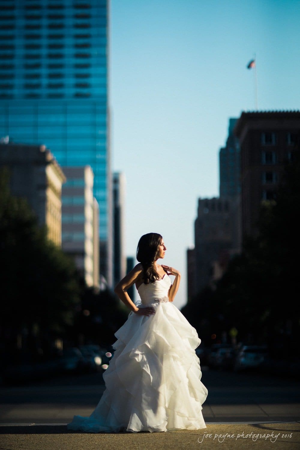 carolina inn wedding photography - kristen & matthew (x2) A woman in a white wedding dress stands confidently in the middle of a city street. The sun is setting, creating a dramatic contrast between the dress and the surrounding skyscrapers. She poses with one hand on her hip, gazing to the side.