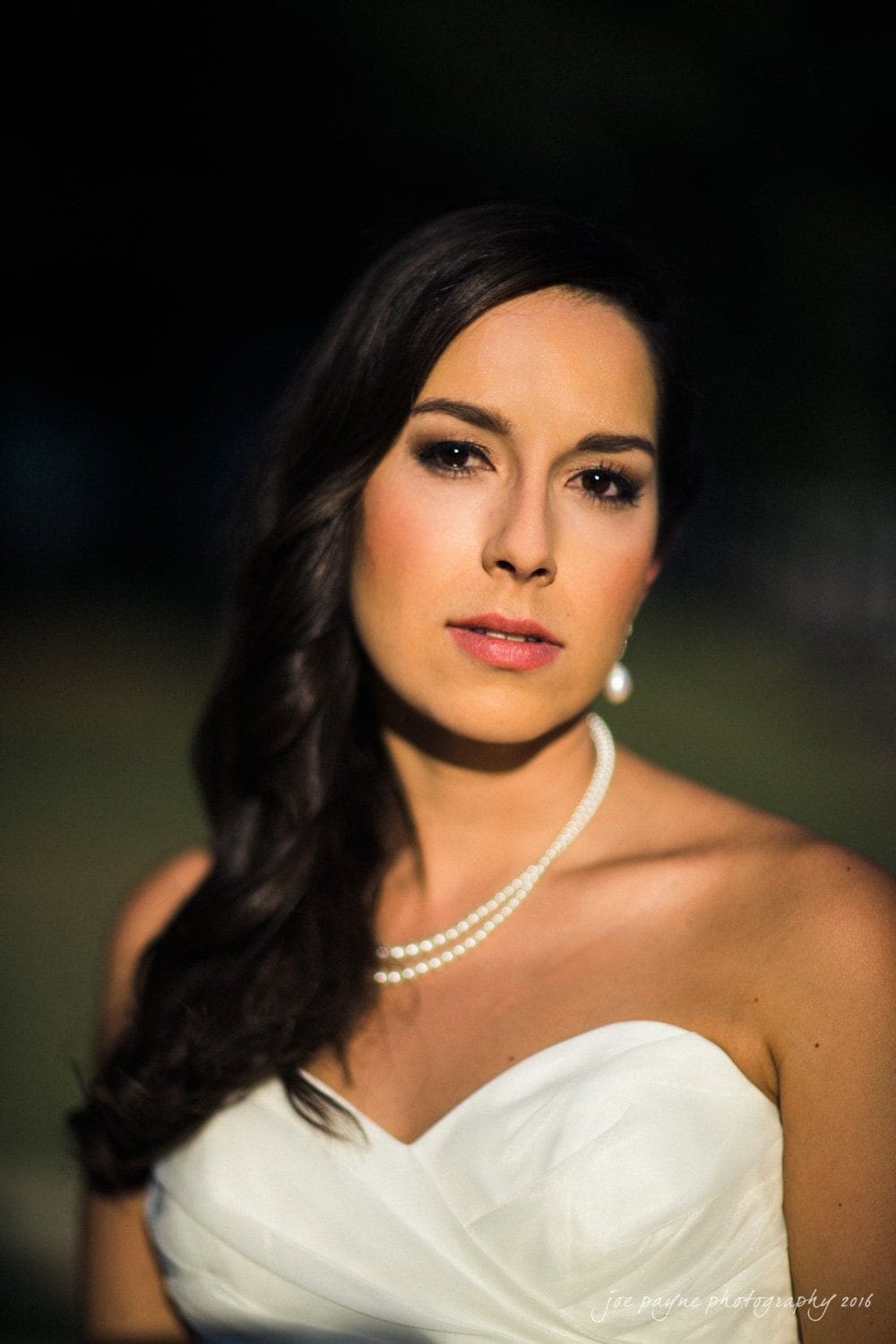 carolina inn wedding photography - kristen & matthew (x2) A woman in a strapless white dress with pearl jewelry stands outdoors. She has long, dark hair styled over one shoulder, and her makeup is softly done. The background is blurred, highlighting her as the main focus.
