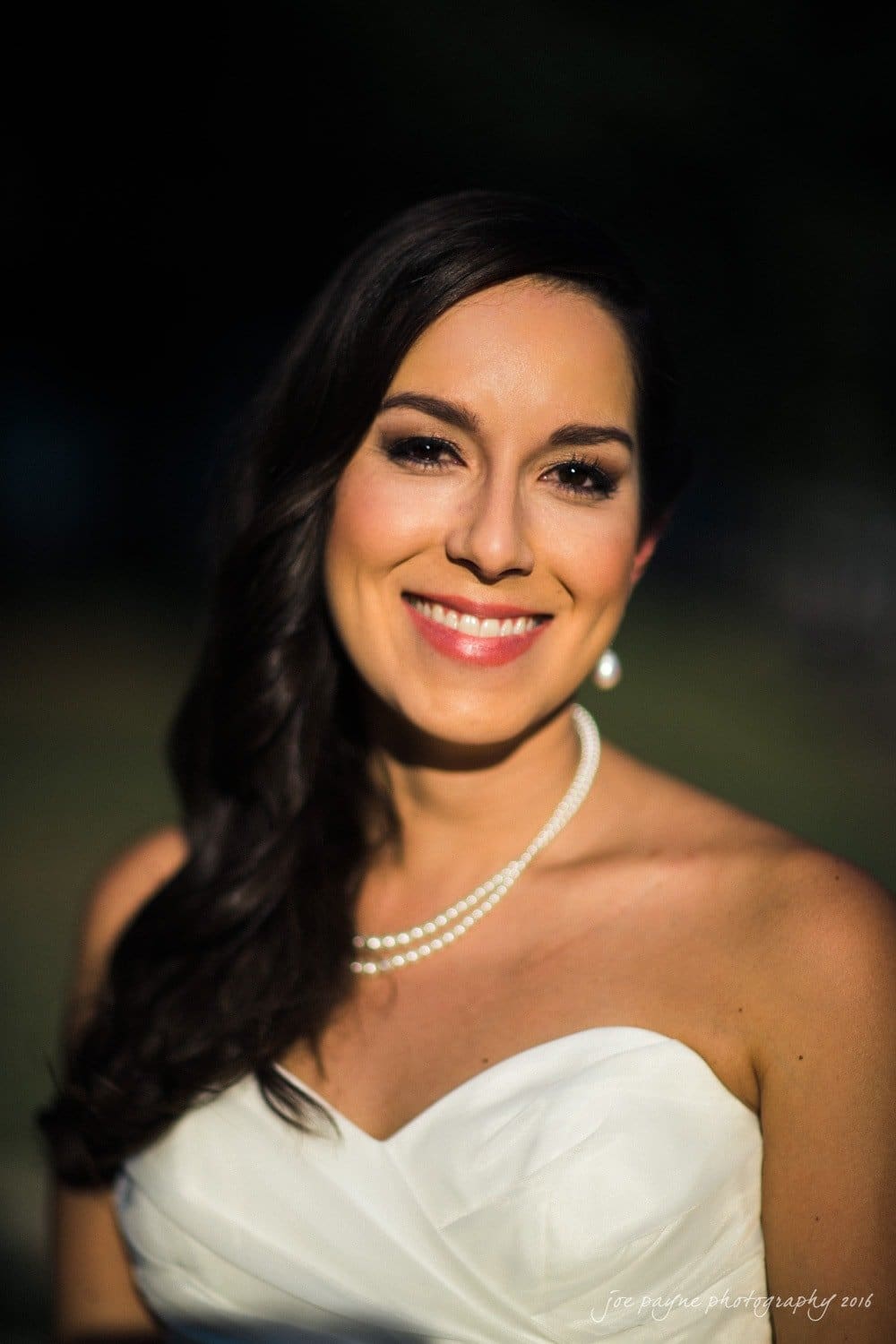 carolina inn wedding photography - kristen & matthew (x2) A woman with long dark hair smiles warmly, wearing a strapless white dress and a pearl necklace. The background is blurred, bringing focus to her radiant expression.