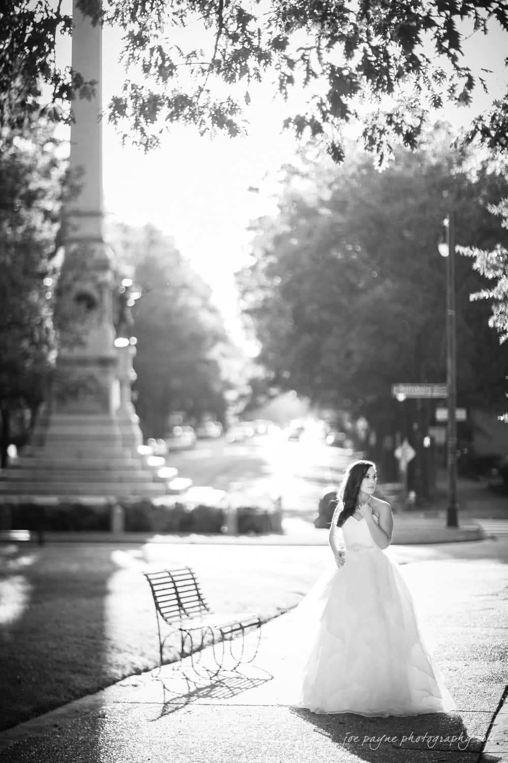 carolina inn wedding photography - kristen & matthew (x2) Black and white photo of a bride standing in a sunlit park, wearing a voluminous wedding dress. A bench and a tall monument are visible in the background, with sunlight creating a glowing effect through the trees.