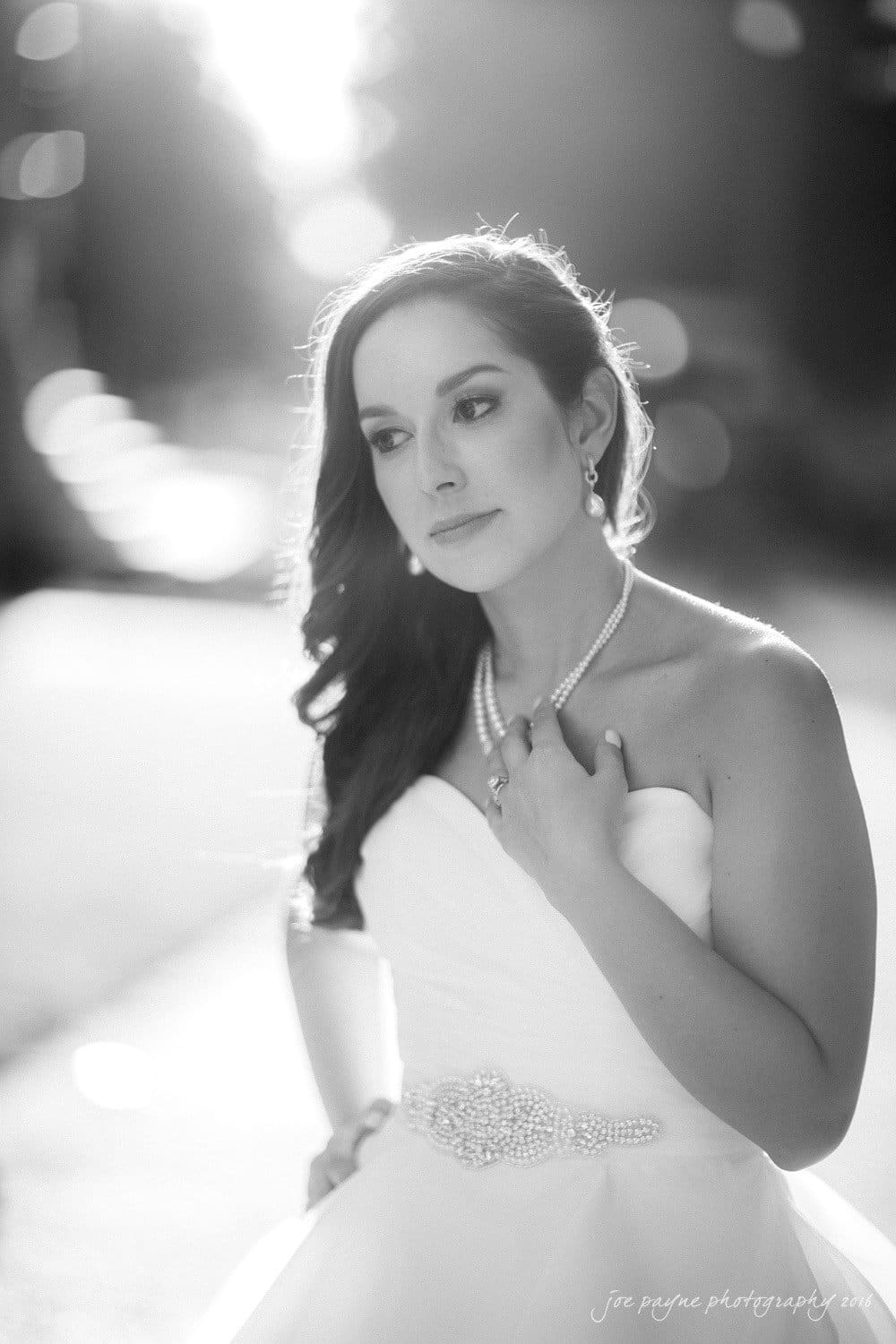 carolina inn wedding photography - kristen & matthew (x2) A grayscale portrait of a woman in a strapless wedding dress, adorned with a pearl necklace and belt. She gazes thoughtfully to the side, standing in soft, dappled sunlight.