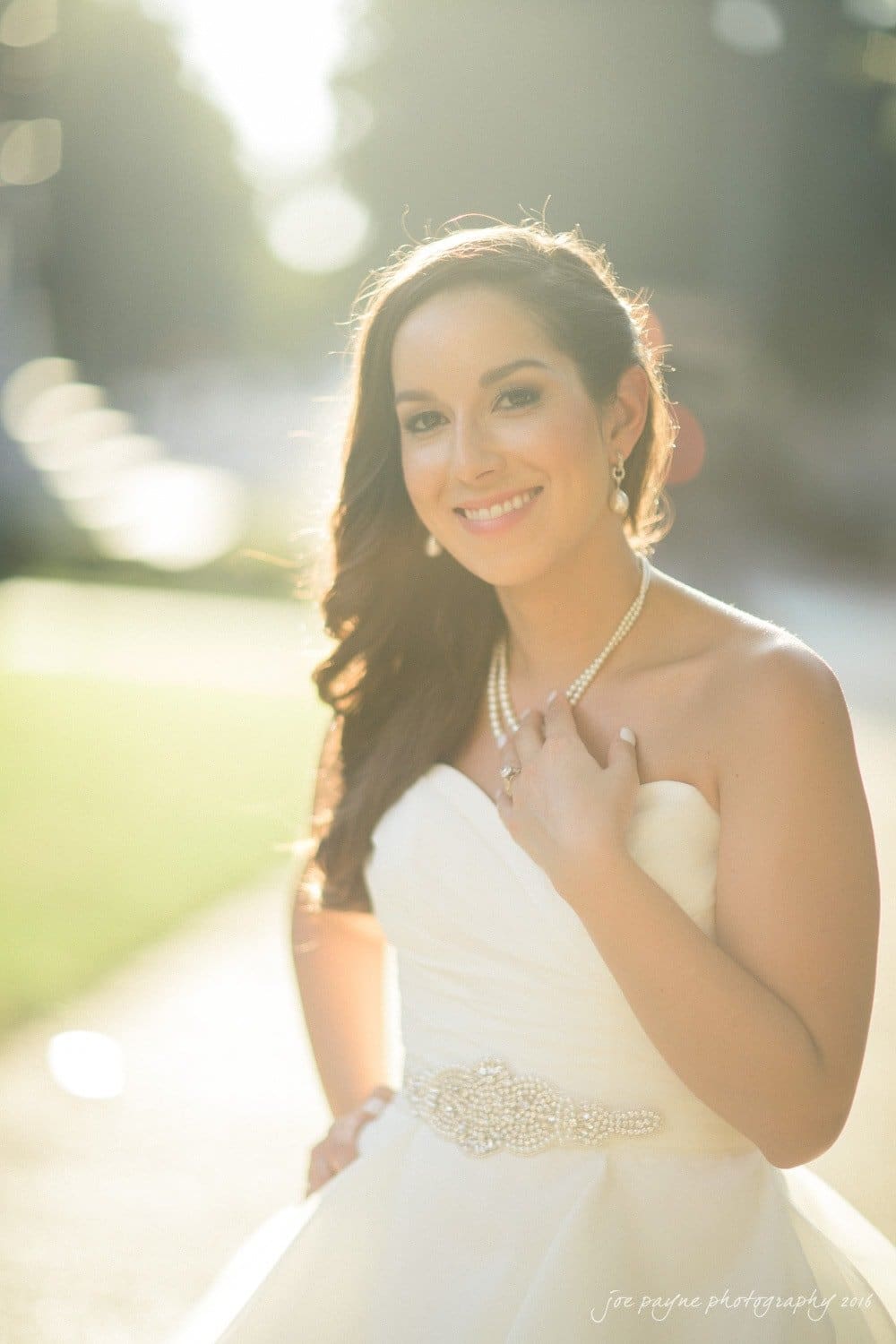carolina inn wedding photography - kristen & matthew (x2) A bride in a strapless wedding dress smiles while standing outdoors. She has long, wavy hair and is wearing pearl jewelry. The background is softly blurred with bright sunlight shining through.