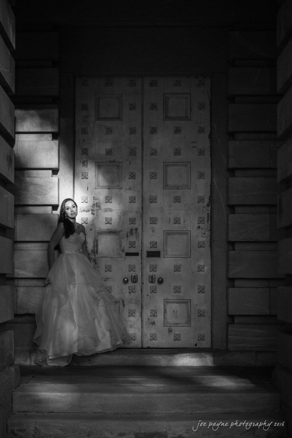 carolina inn wedding photography - kristen & matthew (x2) A woman in a flowing, strapless gown stands in front of a large, ornate door. The scene is in black and white, with dramatic lighting casting shadows across the setting. The image has a serene and contemplative mood.
