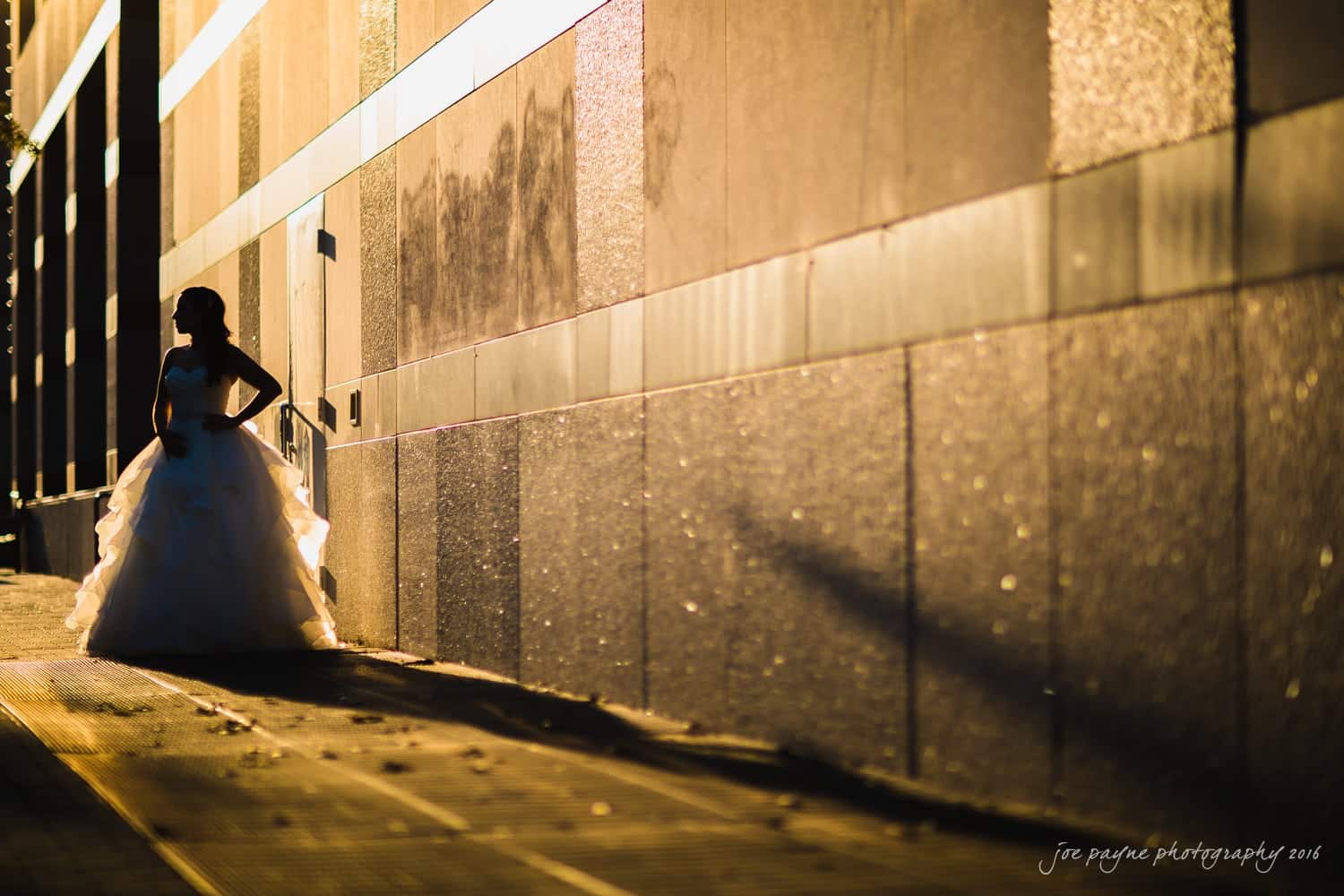 carolina inn wedding photography - kristen & matthew (x2) Silhouetted bride in a flowing gown stands against a sunlit, textured wall. The golden light casts long shadows, creating a dramatic and romantic atmosphere.