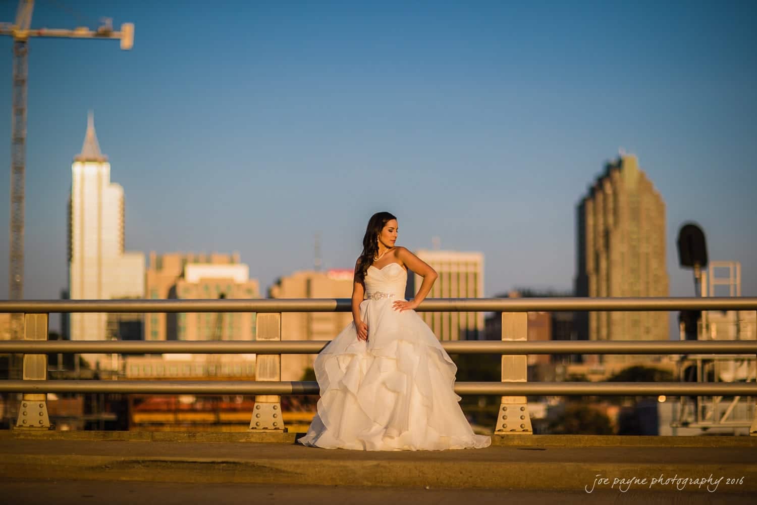 carolina inn wedding photography - kristen & matthew (x2) A woman in a white wedding dress stands confidently on a bridge, with a city skyline and clear blue sky in the background. She is looking to the side, creating an elegant and serene scene.