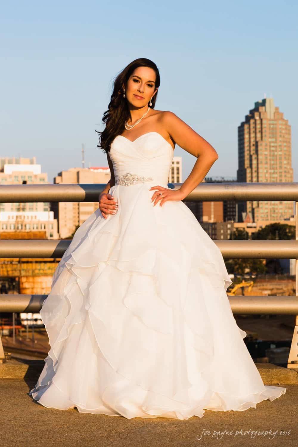 carolina inn wedding photography - kristen & matthew (x2) A woman in a strapless white wedding dress stands on a bridge, with a city skyline in the background. She has long dark hair and wears a necklace, earrings, and a belt with a jeweled embellishment. The setting sun casts a warm glow.