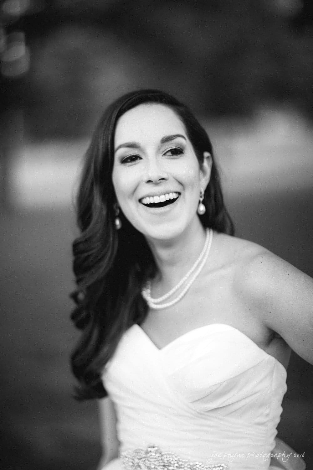 carolina inn wedding photography - kristen & matthew (x2) A woman in a white strapless dress smiles broadly. She has long, dark hair and wears pearl earrings and a pearl necklace. The background is blurred, giving the image a soft focus.