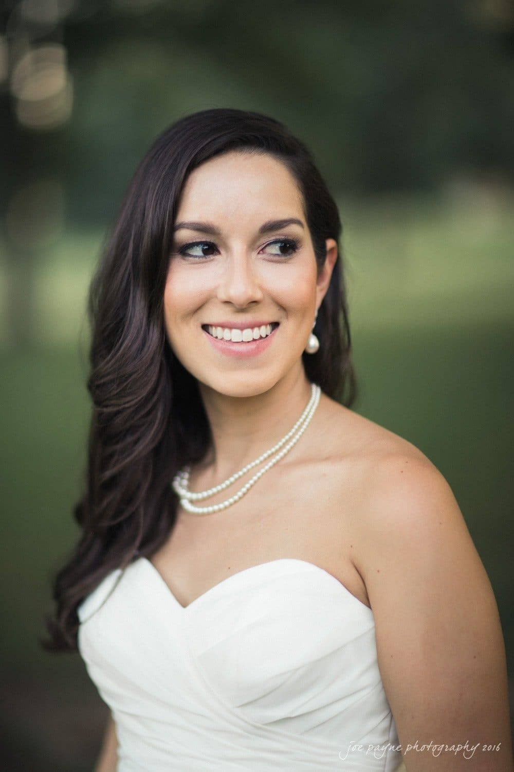 carolina inn wedding photography - kristen & matthew (x2) A bride in a white dress stands outdoors, smiling and looking to the side. She has long dark hair and wears pearl jewelry. The background is blurred greenery.