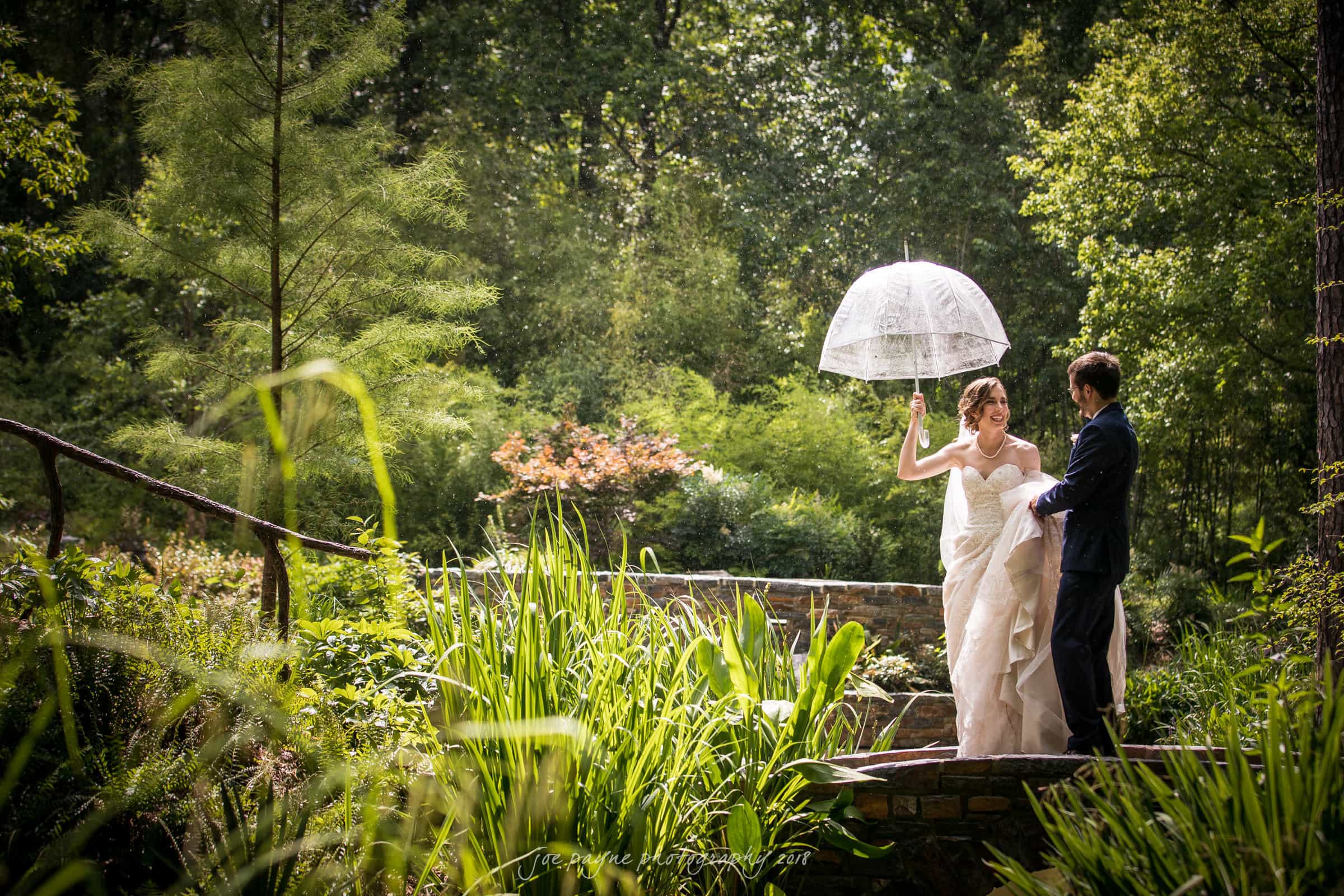 Duke Chapel Weddings A backlit documentary wedding photograph of a bride and groom on the bridge at Welch Woodland Garden Overlook in Sarah P. Duke Gardens, smiling after a rainstorm as the groom holds her dress.