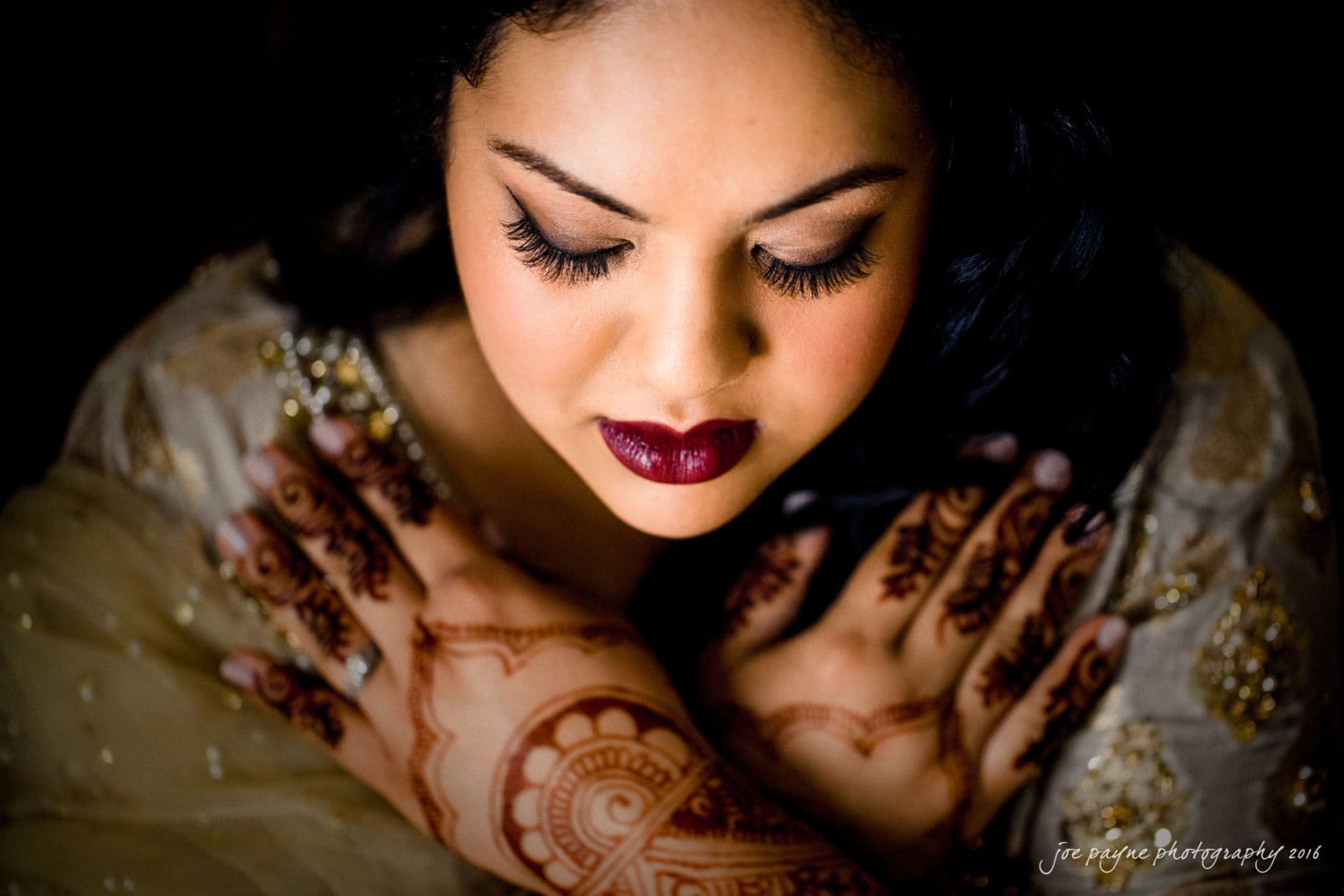 A woman with deep red lipstick and dramatic eye makeup looks down, crossing her hands over her chest, henna designs visible. Her gold-embellished cream garment evokes the elegance of Prestonwood Country Club weddings.