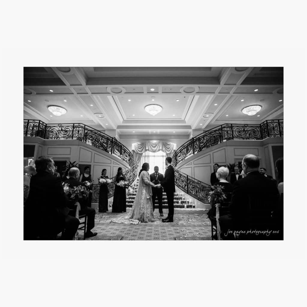 A black and white photo of a wedding ceremony taking place indoors. The couple stands facing each other, holding hands. Guests are seated on both sides, and a grand staircase with ornate railings serves as the backdrop. Elegant chandeliers hang from the ceiling.